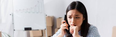 Scared asian volunteer talking on telephone with blurred charity center on background, banner clipart