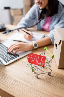 Close up view of price tag with sale lettering in shopping cart near carton box on desk with blurred woman on background clipart
