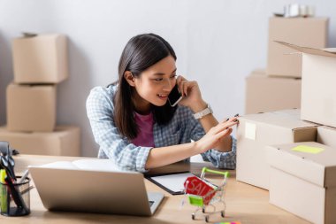 Gülümseyen Asyalı gönüllü akıllı telefondan konuşuyor. Yardım merkezinde, bulanık arka plandaki karton kutuya dokunurken.