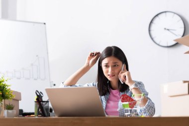 Korkak Asyalı gönüllü akıllı telefondan konuşuyor. Bulanık ön planda, hayır kurumunda karton kutuların yanında dizüstü bilgisayara bakıyor.