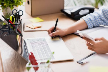 Masasındaki etikette akıllı telefon yazısıyla, kırtasiye malzemesi ve bulanık ön planda cihazları olan kadının kırpılmış görünümü