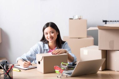 Mutlu Asyalı gönüllü, karton kutusuyla kameraya bakıyor. Masadaki panoya yazıyor. Yardım merkezinde, bulanık arka planda.