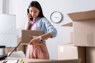 Yüzünde paketle gülümseyen Asyalı gönüllü telefonda konuşurken yardım merkezindeki karton kutuların yanında bulanık ön planda duruyor.