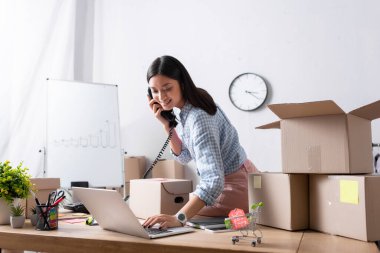 Gülümseyen Asyalı gönüllü, yardım merkezinde karton kutuların yanında otururken telefonla konuşuyor ve dizüstü bilgisayarla yazı yazıyor.