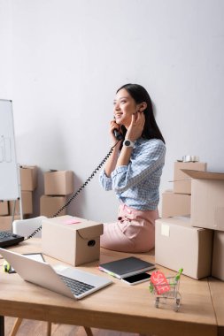 Gülümseyen Asyalı gönüllü, yardım merkezinde masasında cihazlar ve karton kutularla otururken telefonla konuşuyor.