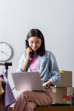 Ofisteki kutuların ve kıyafetlerin yanında dizüstü bilgisayar kullanırken telefonla konuşan neşeli Asyalı gönüllü. 