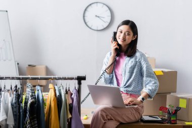 Gülümseyen Asyalı gönüllü telefonla konuşuyor ve yardım merkezinde kıyafet ve karton kutuların yanında dizüstü bilgisayar kullanıyor. 