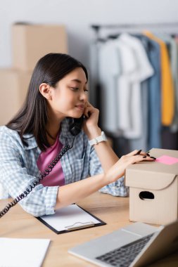 Asyalı gönüllü telefonda pano, kutu ve dizüstü bilgisayarın yanında konuşuyor. 