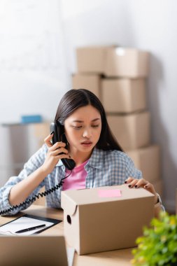 Bağış merkezinin Asyalı yöneticisi telefon başında pano, kutu ve dizüstü bilgisayar başında bulanık ön planda konuşuyor 