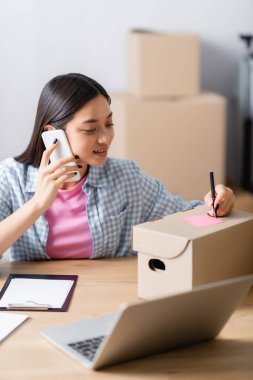 Gülümseyen Asyalı gönüllü akıllı telefondan konuşuyor. Önplanda bulanık dizüstü bilgisayar ve pano yanında yapışkan notla kutunun üzerine yazı yazıyor.