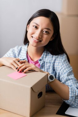 Smiling asian woman looking at camera near box with sticky note in charity center  clipart