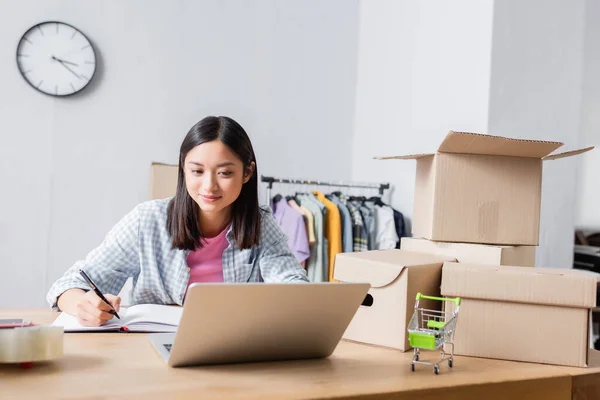 Smiling Asian Volunteer Looking Laptop While Writing Notebook Desk Carton — Stock Photo, Image