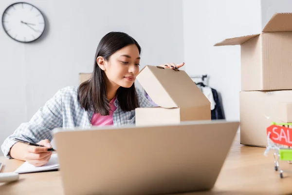 Positive Asian Volunteer Opening Looking Carton Box While Sitting Desk — Stock Photo, Image