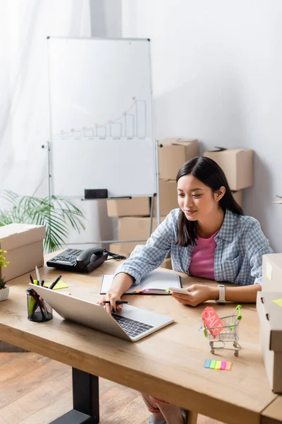 Smiling Asian Volunteer Smartphone Typing Laptop While Sitting Desk Carton — Stok Foto