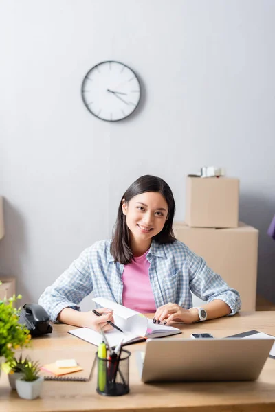 Smiling Asian Volunteer Looking Camera While Leafing Notebook Desk Devices — Stok Foto