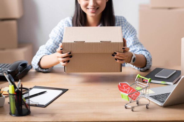 Cropped view of woman holding carton box while sitting at desk with price tag in shopping cart on blurred background
