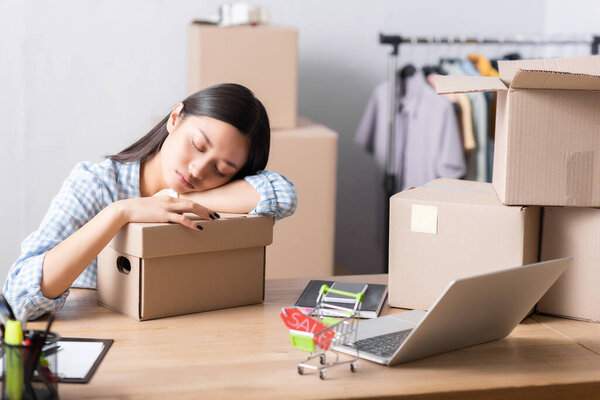 Tired asian volunteer sleeping on carton box while sitting at desk with blurred packages and hanging rack on background