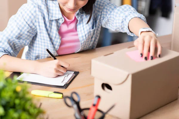 Cropped view of female volunteer writing on clipboard while holding carton box at desk on blurred foreground