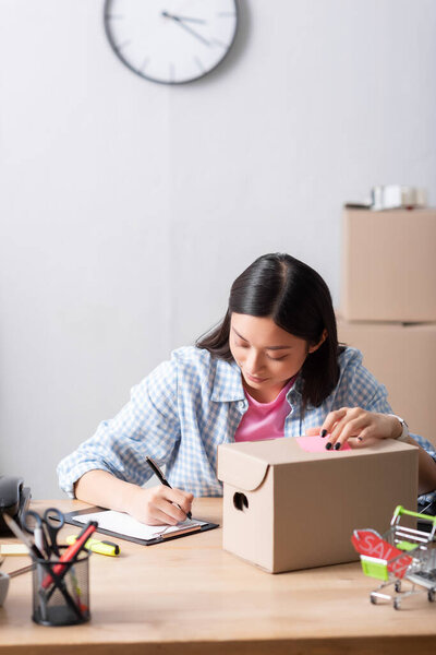 Positive asian volunteer writing on clipboard while sitting at desk with carton box in charity center on blurred background