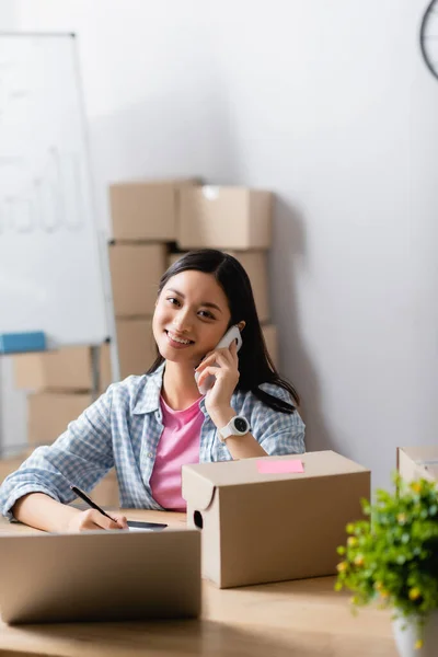 Smiling Asian Volunteer Talking Smartphone Carton Boxes Laptop Blurred Foreground — Stok Foto