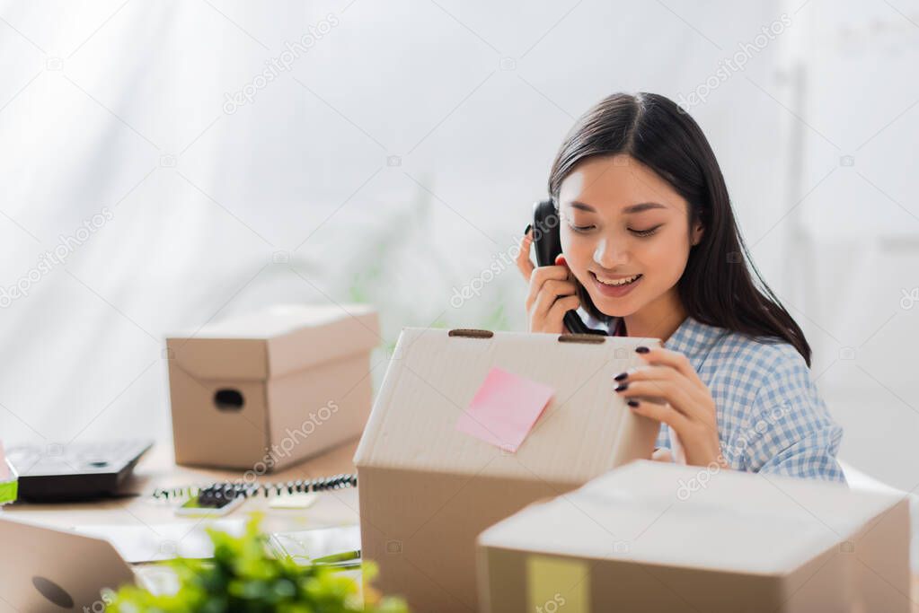 Smiling asian volunteer talking on telephone and looking at cardboard box on blurred foreground 
