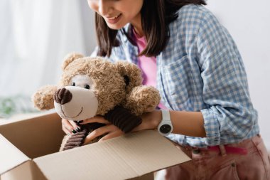 Cropped view of volunteer smiling while putting soft toy in box in charity office  clipart