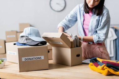 Cropped view of asian woman holding clothes near boxes with donations lettering and caps in charity center  clipart