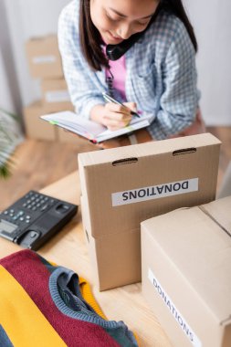 Smiling asian woman talking on telephone and writing on notebook near boxes with donations and clothes  clipart