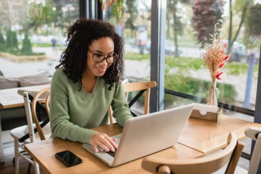 african american freelancer in glasses using laptop in cafe  clipart