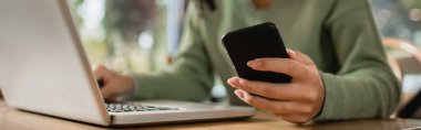Masanın üzerinde dizüstü bilgisayarın yanında telefonu olan Afro-Amerikan kadının kısmi görüntüsü 