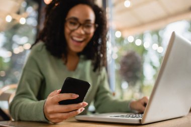 Akıllı telefon, arka planda bulanık gözlüklü heyecanlı Afrikalı Amerikalı kadının elinde. 