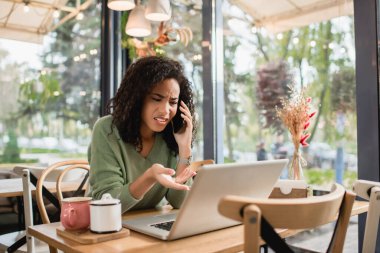 Afro-Amerikalı hoşnutsuz bir kadın akıllı telefondan konuşuyor ve kafede dizüstü bilgisayarını işaret ediyor.