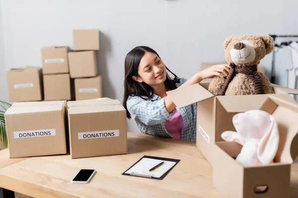 Smiling asian volunteer putting soft toy in package near smartphone and clipboard on table