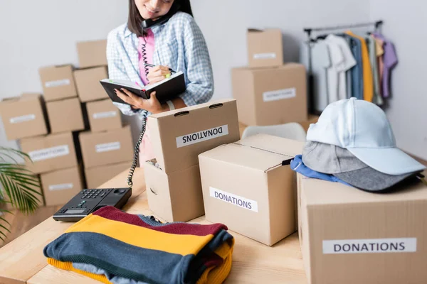 Cropped view of smiling volunteer talking on telephone and writing on notebook near clothes and boxes with donations lettering