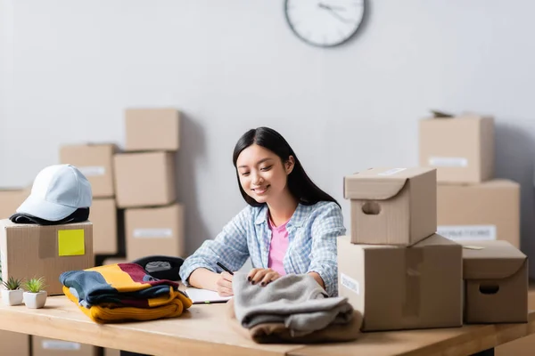 Cheerful Asian Volunteer Holding Clothes Writing Notebook Cardboard Boxes Blurred — Stock Photo, Image