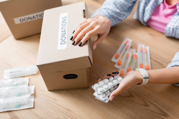 Обрезанный вид женщины, держащей таблетки рядом со шприцами и коробкой с надписями пожертвований на столе 