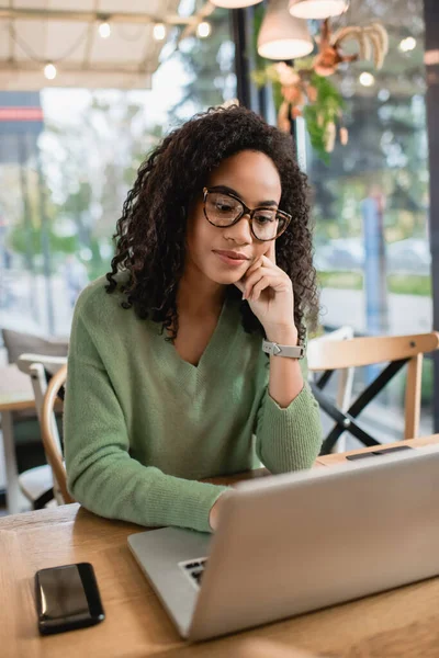 Mujer Afroamericana Gafas Usando Portátil Cerca Teléfono Inteligente Cafetería —  Fotos de Stock