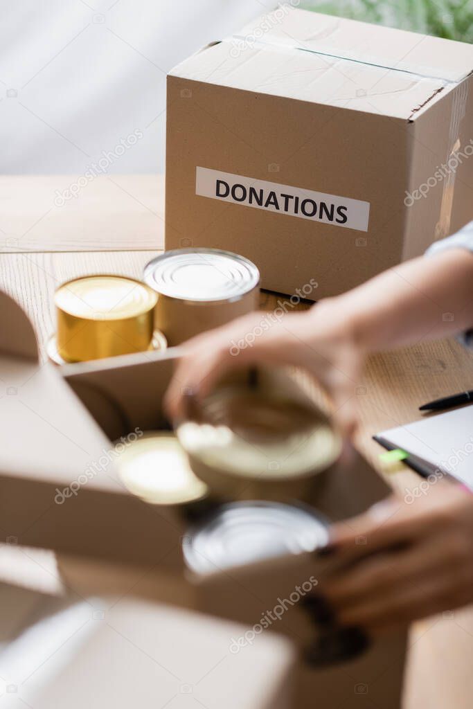 Cropped view of package with donations lettering near tin cans and volunteer on blurred foreground 