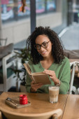 happy african american woman in eyeglasses reading book near glass with latte