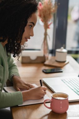 african american woman writing in notebook near cup of latte while studying online in cafe clipart