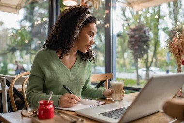 Afro-Amerikan kadın kulaklıkla podcast dinliyor ve kahve bardağıyla kafeteryada laptopun yanında kahve tutuyor. 