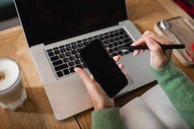 Afro-Amerikan kadının elinde bilgisayarın yanında boş ekranlı akıllı telefon, bir bardak latte ve defter tutuşu. 