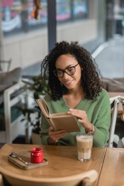 happy african american woman in eyeglasses reading book near glass with latte clipart
