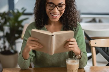 Neşeli Afro-Amerikalı kadın, kafeteryada kahve içip kitap okuyor.
