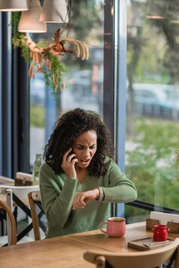 Afro-Amerikan kadın, bilek kontrolü yaparken akıllı telefondan konuşuyor ve kafede bekliyor.