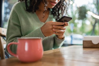 Afrika kökenli Amerikalı bir kadının ön planda bulanık bir fincanın yanında akıllı telefondan mesajlaşması görüntüsü. 