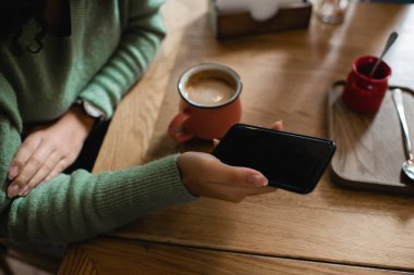 Kapuçino bardağının yanında elinde boş ekranla akıllı telefon tutan Afro-Amerikan kadının kısmi görüntüsü 