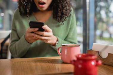 Afro-Amerikan bir kadının ön planda bulanık bir şekilde akıllı telefondan mesaj atmasına çok az kalmış bir fincan kahve. 