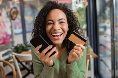 Neşeli Afrikalı Amerikalı kadın elinde akıllı telefon ve kredi kartıyla gülüyor.