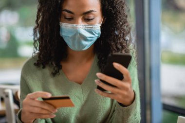 Tıbbi maskeli kıvırcık Afrikalı Amerikalı kadın kafeteryada akıllı telefon ve kredi kartı tutuyor.